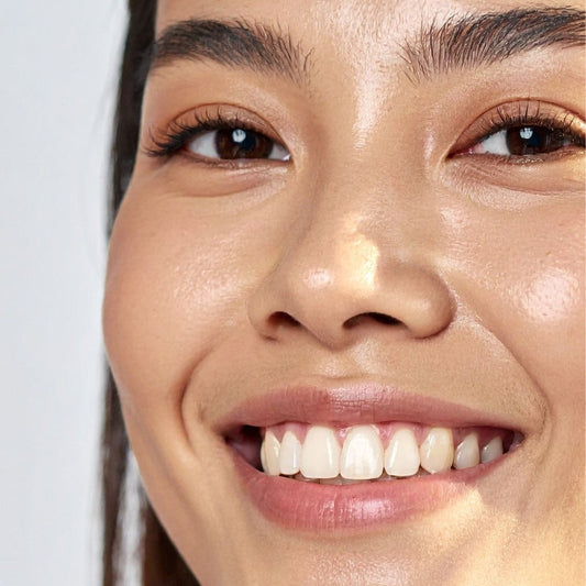 Close-up of a smiling woman's face showing bright, even skin and white teeth against a light gray background.